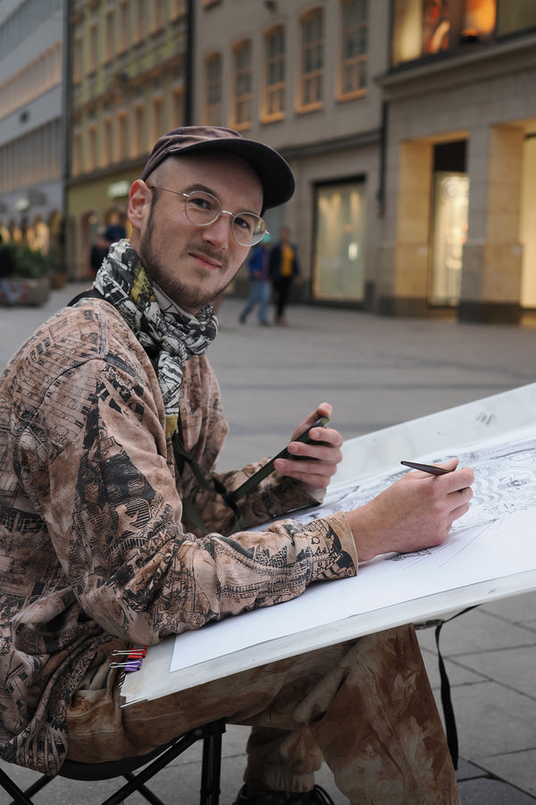 Heiner Radau bei der Arbeit am Zeichenbrett — das detailreiche Muenchen-Stadtkunstwerk entsteht live vor Ort mit Tinte und Feder.