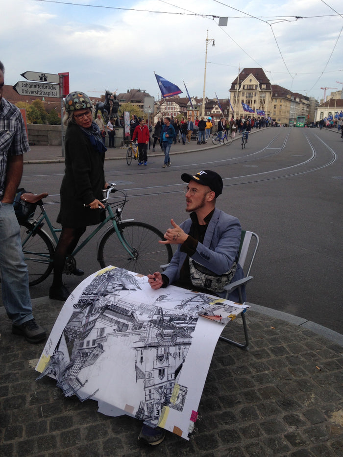 Heiner Radau drawing Basel on the street, engaging with locals as he explains his hand-drawn black-and-white fountain pen portrait collage of the city, featuring bridges, old monuments, and other iconic landmarks